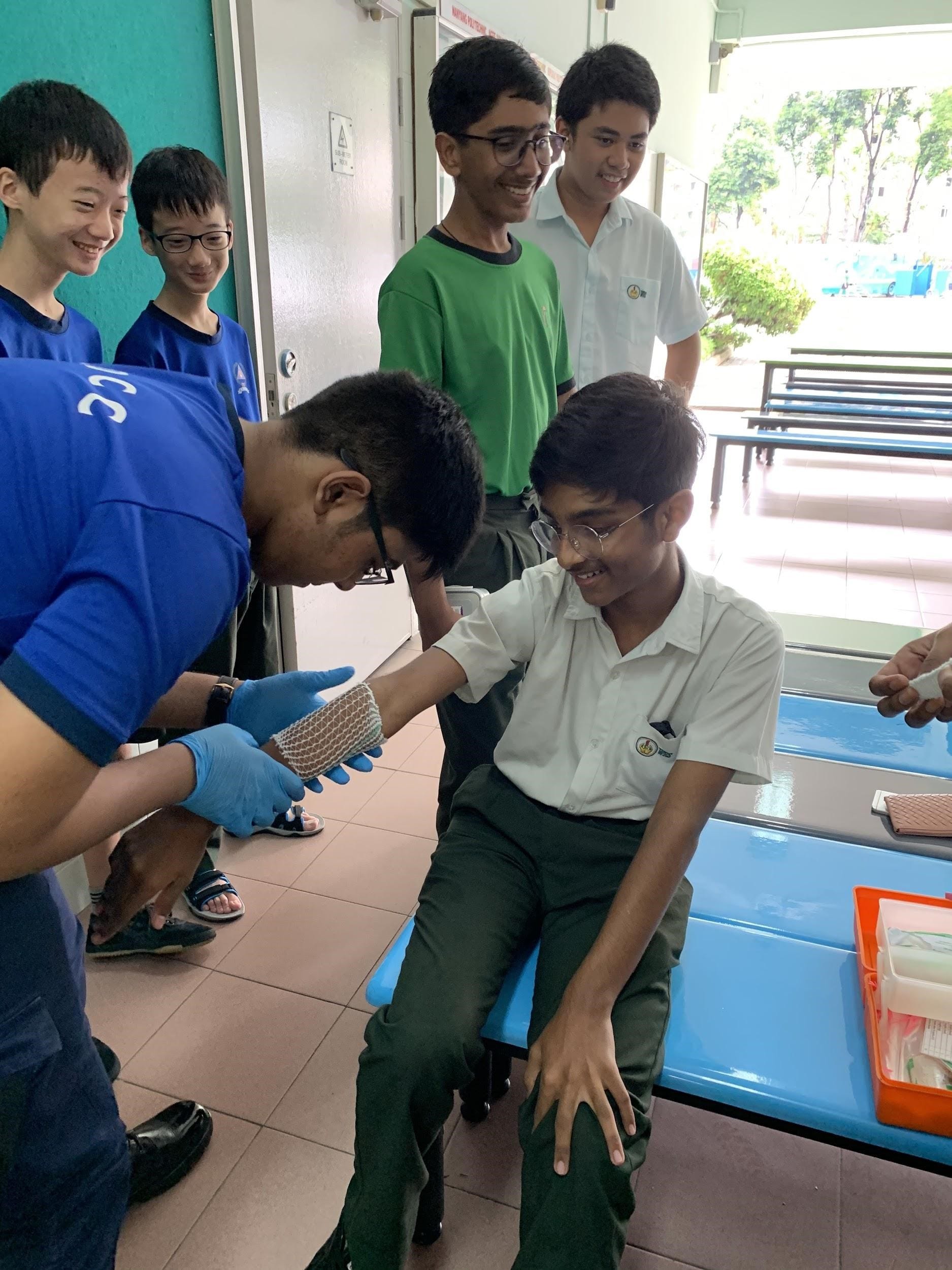 A student wearing gloves applies a bandage to a seated boy's hand while four other students watch and smile.